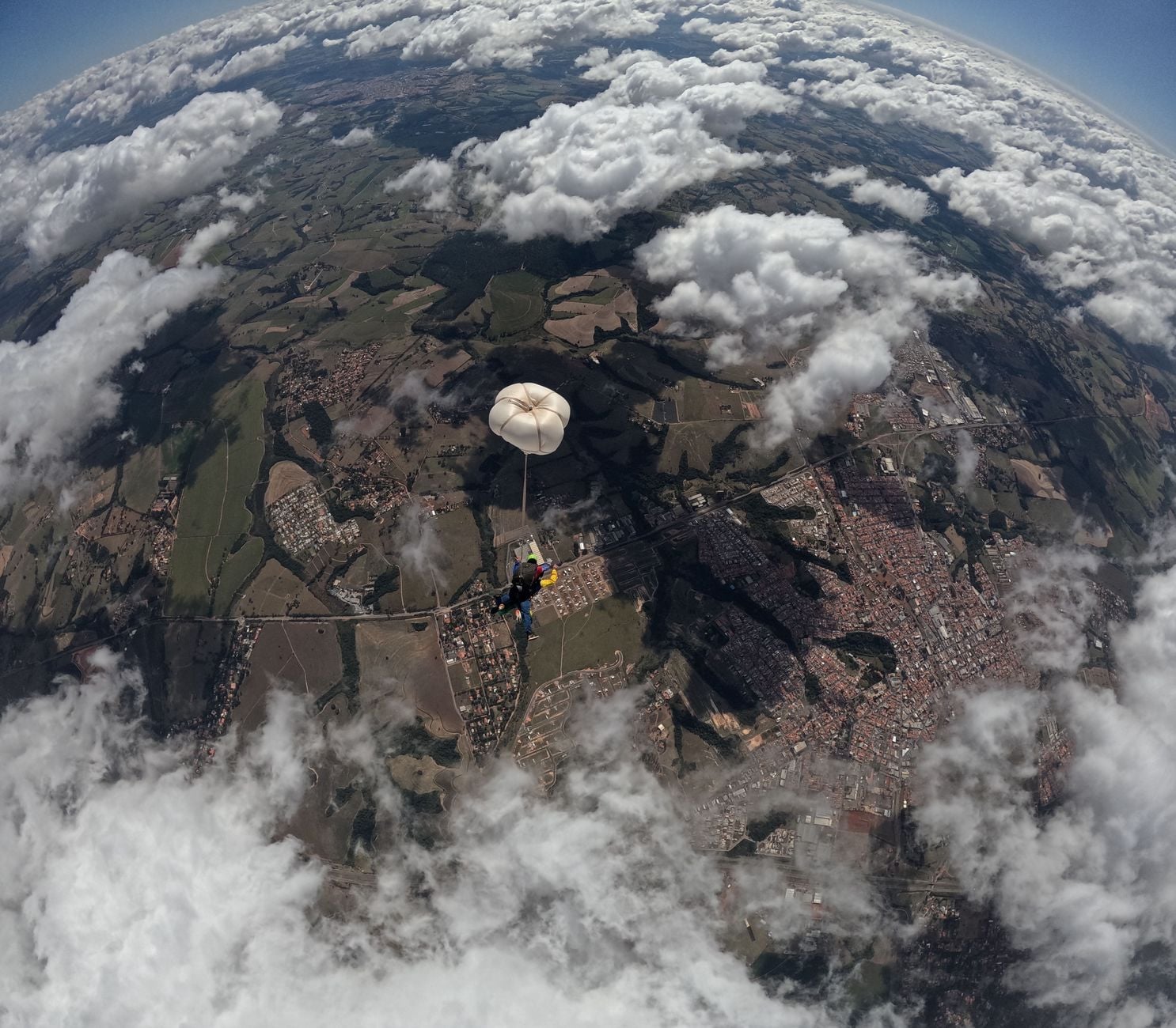 Skydiver w białym spadochronie podczas zeskoku, widok z góry na tereny zurbanizowane i pola odsłonięte przez przerwy w chmurach