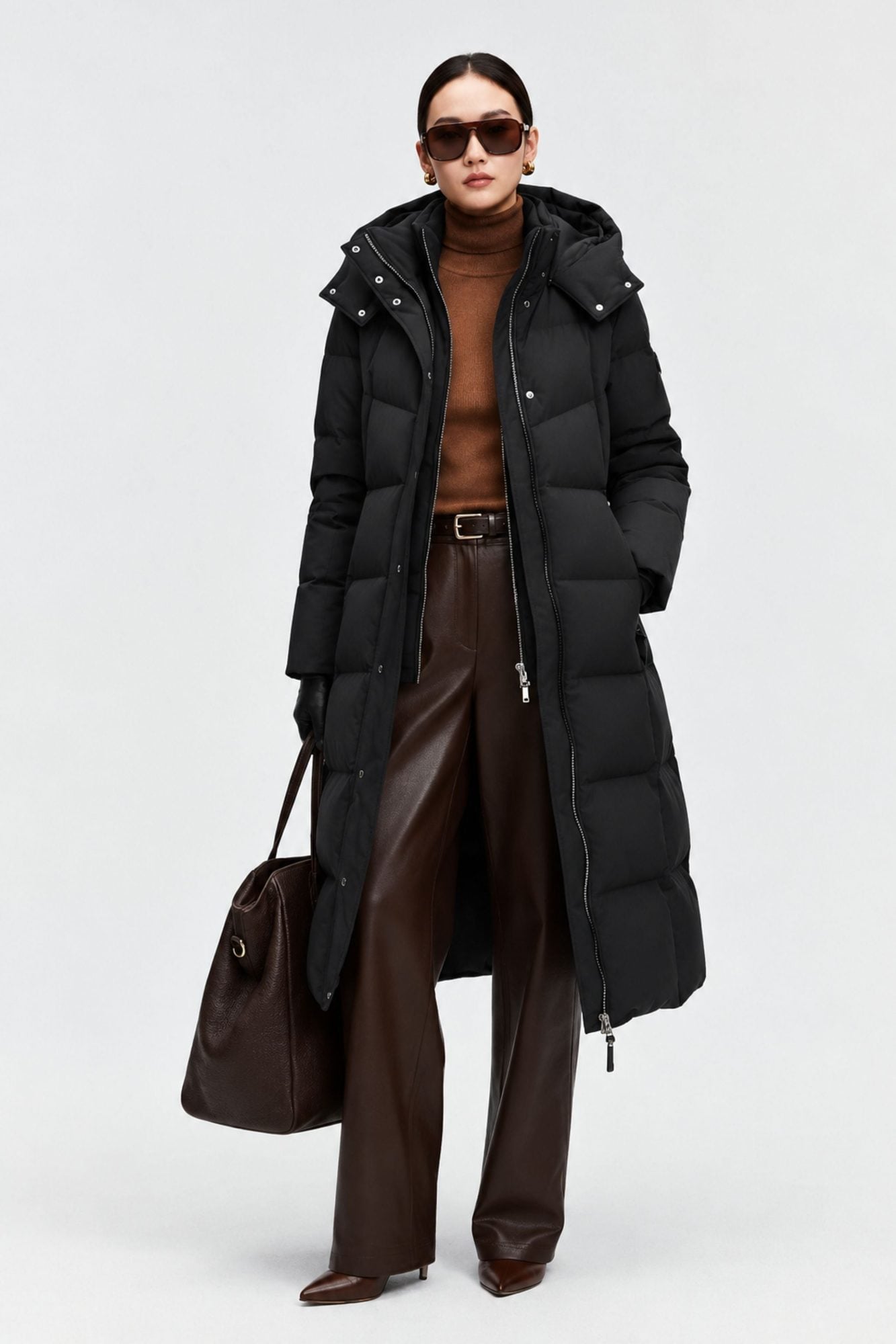 Woman wearing black puffer coat, brown sweater, brown leather pants, sunglasses, and carrying brown bag