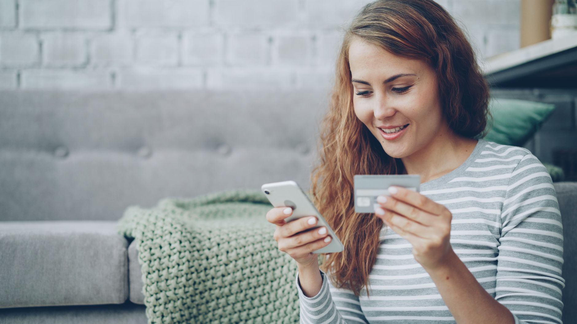 Kobieta siedząca na kanapie trzymająca smartfon i kartę kredytową, uśmiechająca się podczas robienia zakupów online
