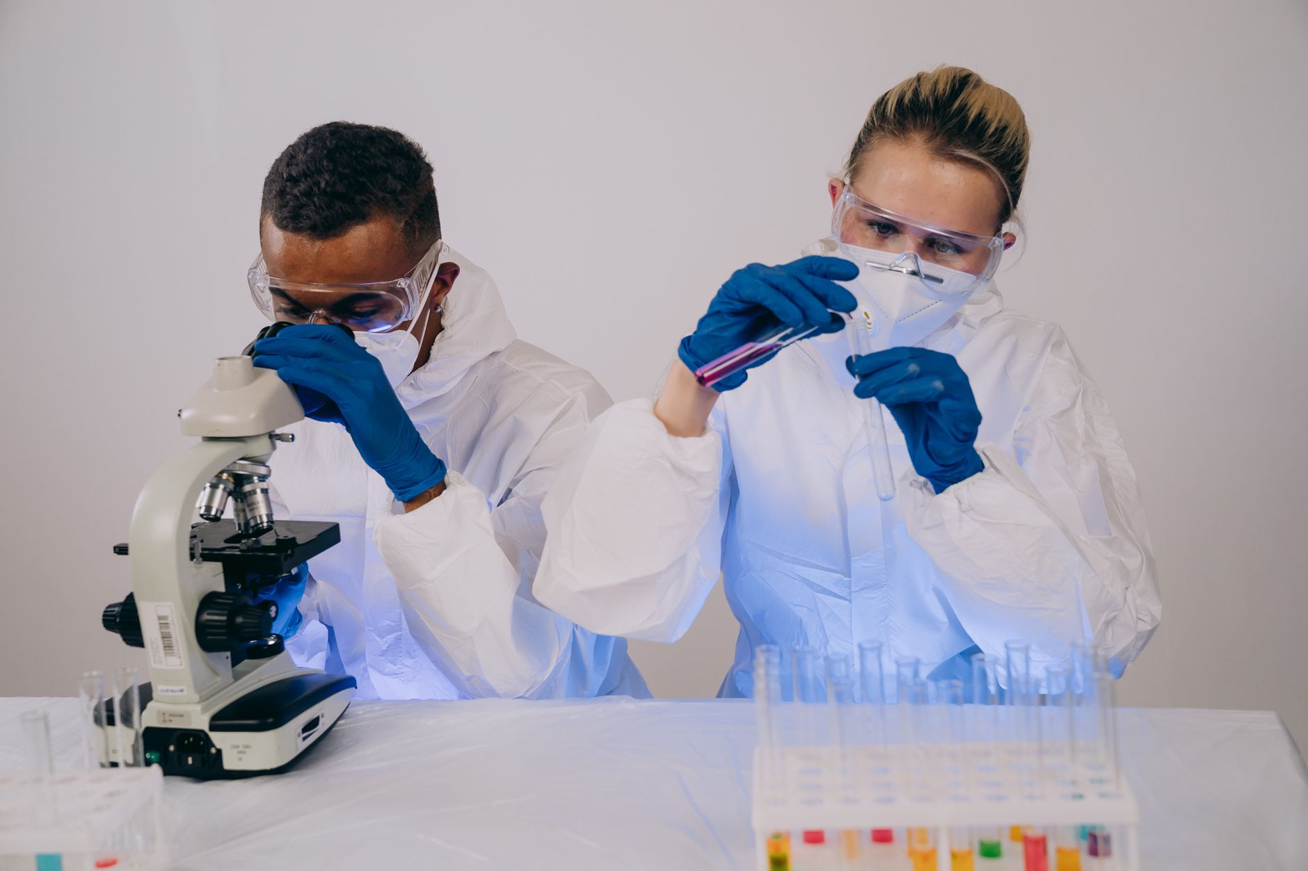 Dwaj naukowcy w białych fartuchach i niebieskich rękawiczkach pracują z mikroskopem i probówkami zawierającymi próbki do badań laboratoryjnych.
